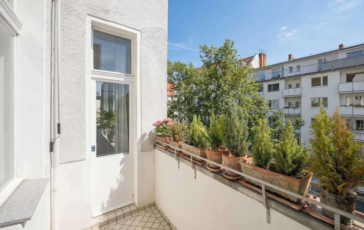A white balcony with potted plants and a view of trees and buildings.
