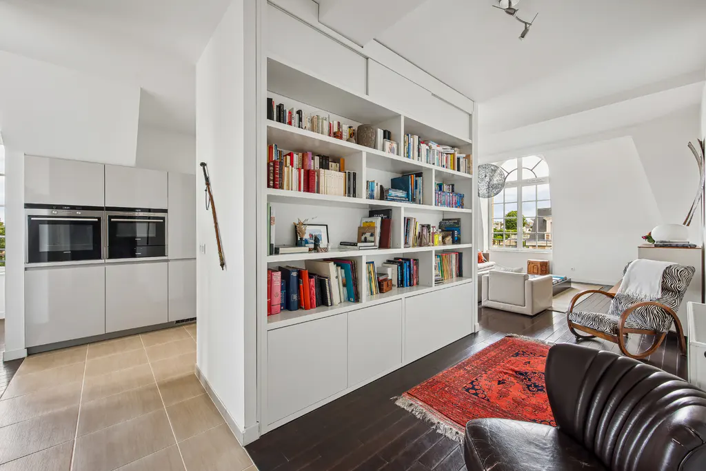 Modern living room with a large white bookshelf, a red patterned rug, a zebra-print armchair, and an adjacent kitchen.