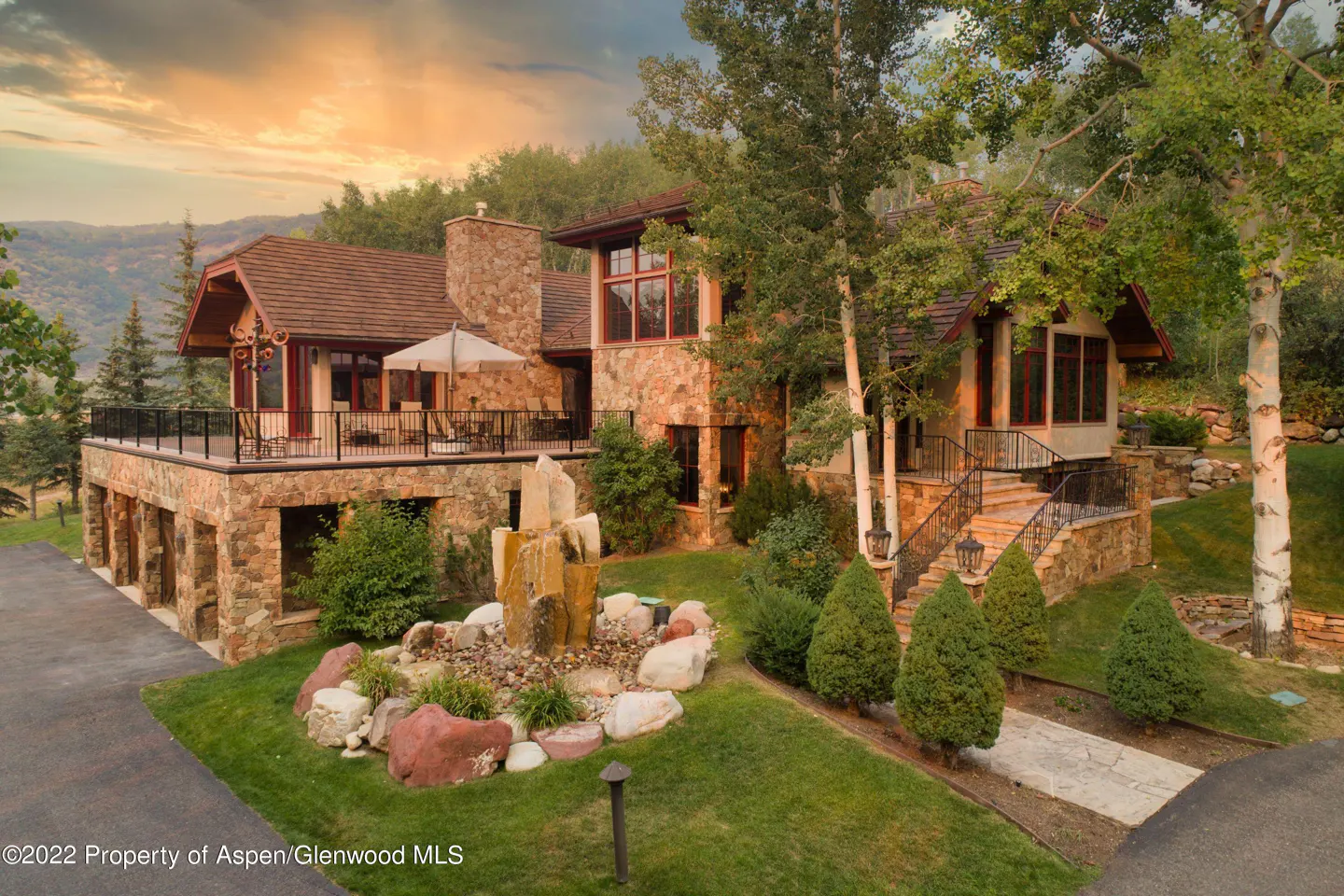 Exterior view of a stone house with a water feature, a deck with furniture, and a mountain backdrop.