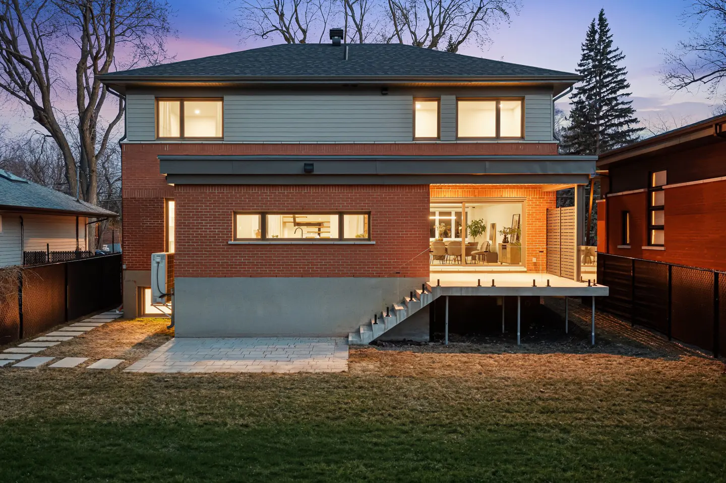 Back of a two-story brick house with a gray roof and siding, a deck, and a green lawn at dusk.