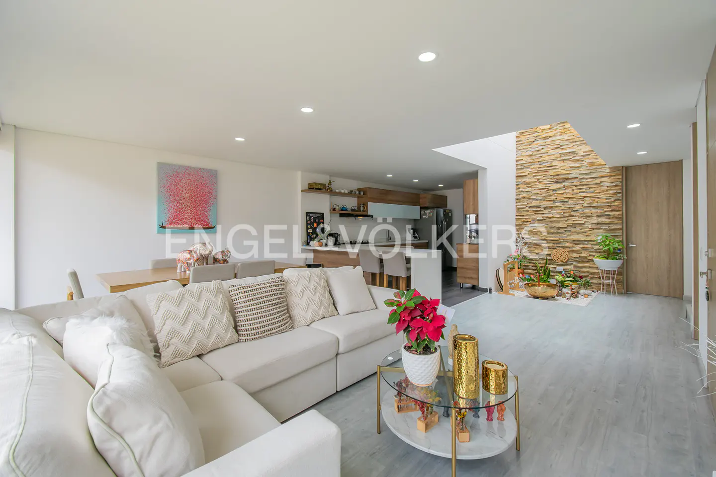 Bright, modern living room with a white sectional sofa, glass coffee table, and stone accent wall. Kitchen and dining area visible in the background.
