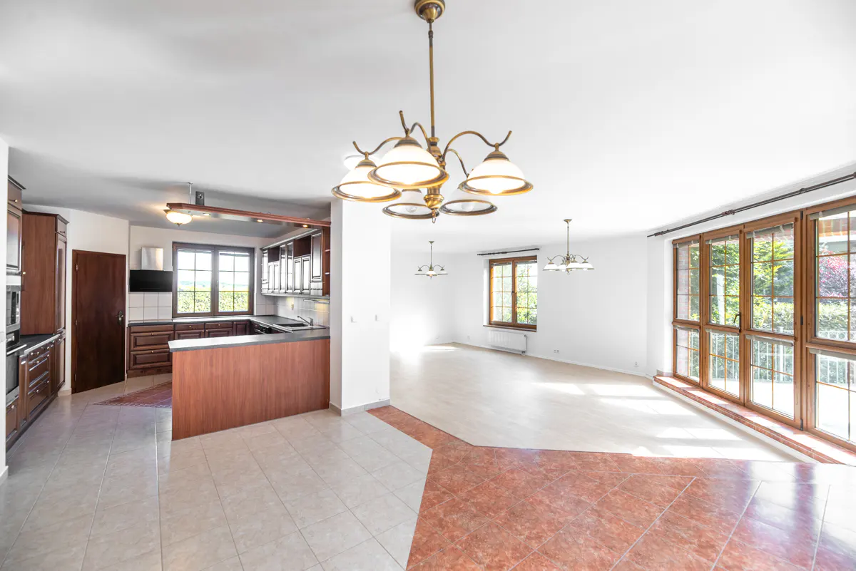 Open-concept kitchen and living room with brown cabinets, tile floors, and large windows. A chandelier hangs from the white ceiling.