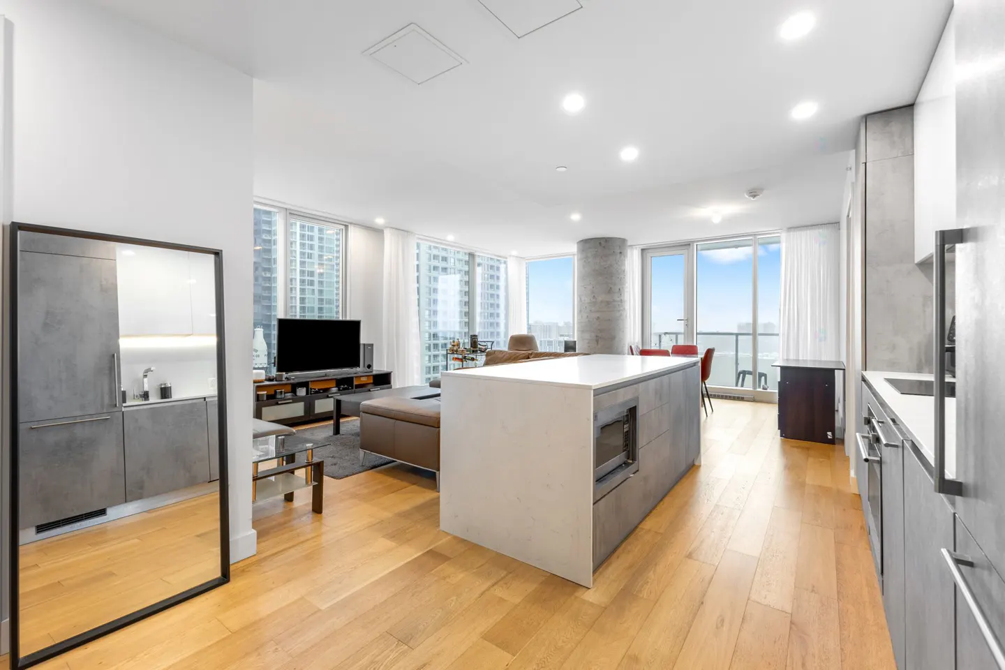 Modern open-concept apartment with a white kitchen island, grey cabinetry, hardwood floors, and a living area with city views.