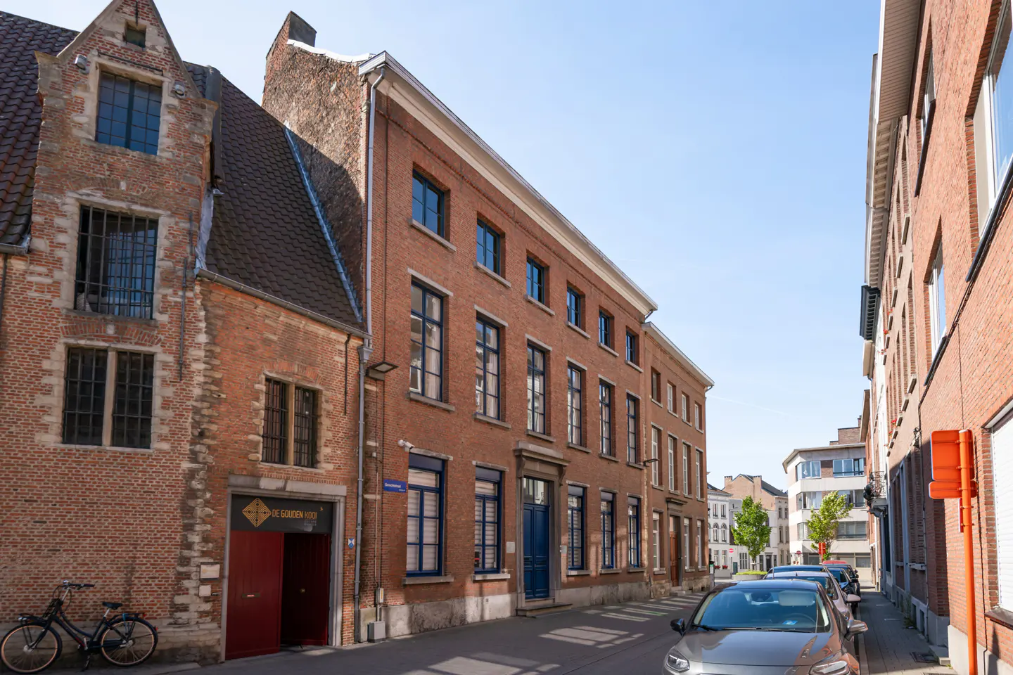 Street view of red brick buildings with blue doors and windows, parked cars, and a bicycle.