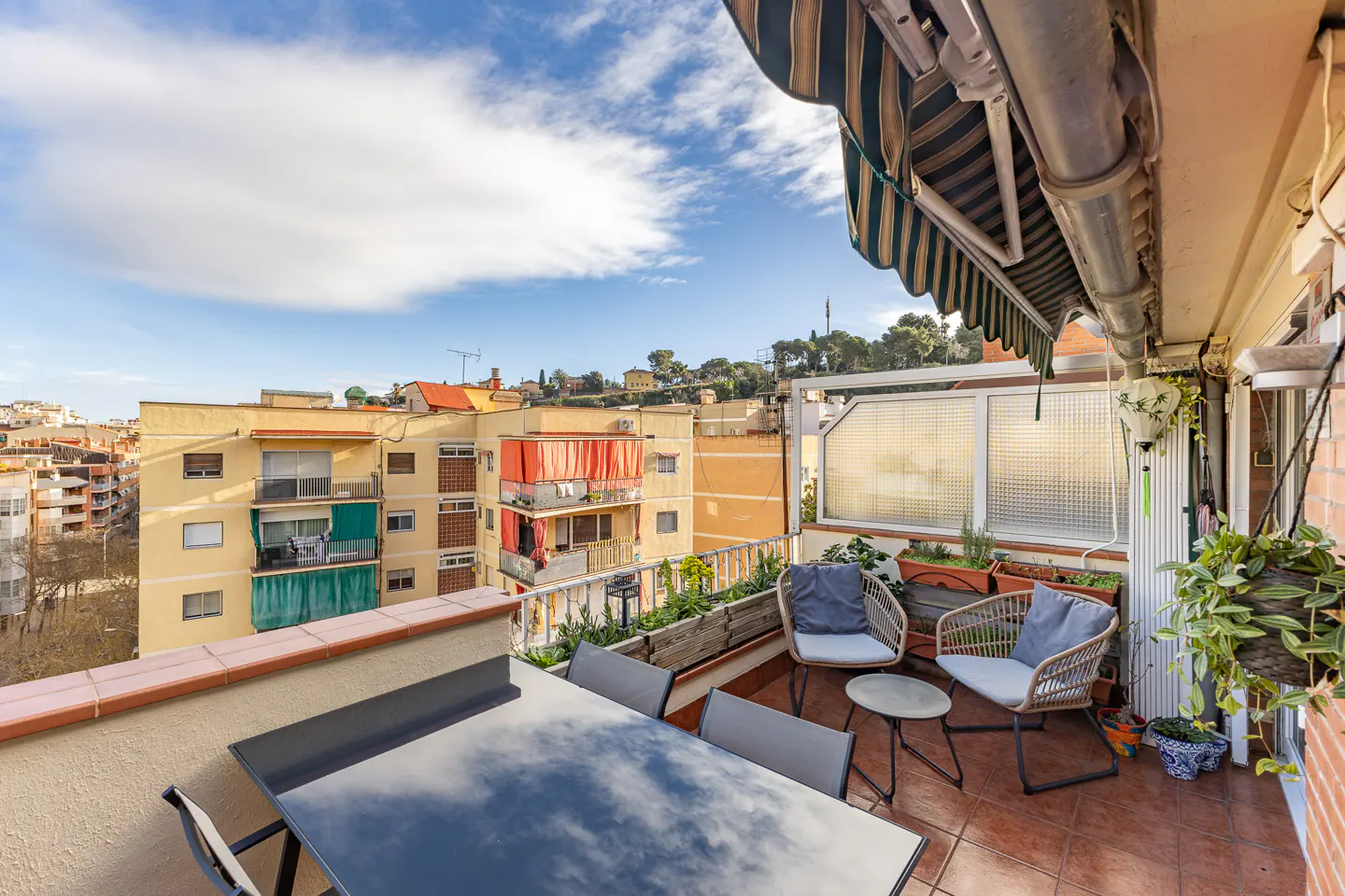 A balcony with a table, chairs, and plants overlooks a city with a blue sky and clouds.