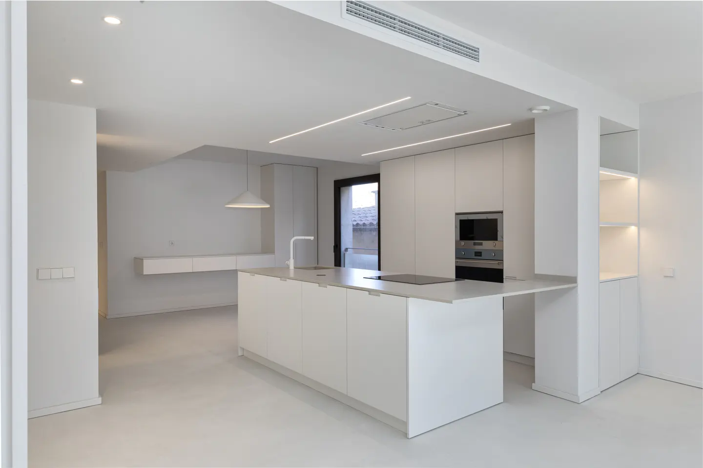 A modern, all-white kitchen with an island, sink, and built-in oven. Open shelving and recessed lighting add to the bright, minimalist design.