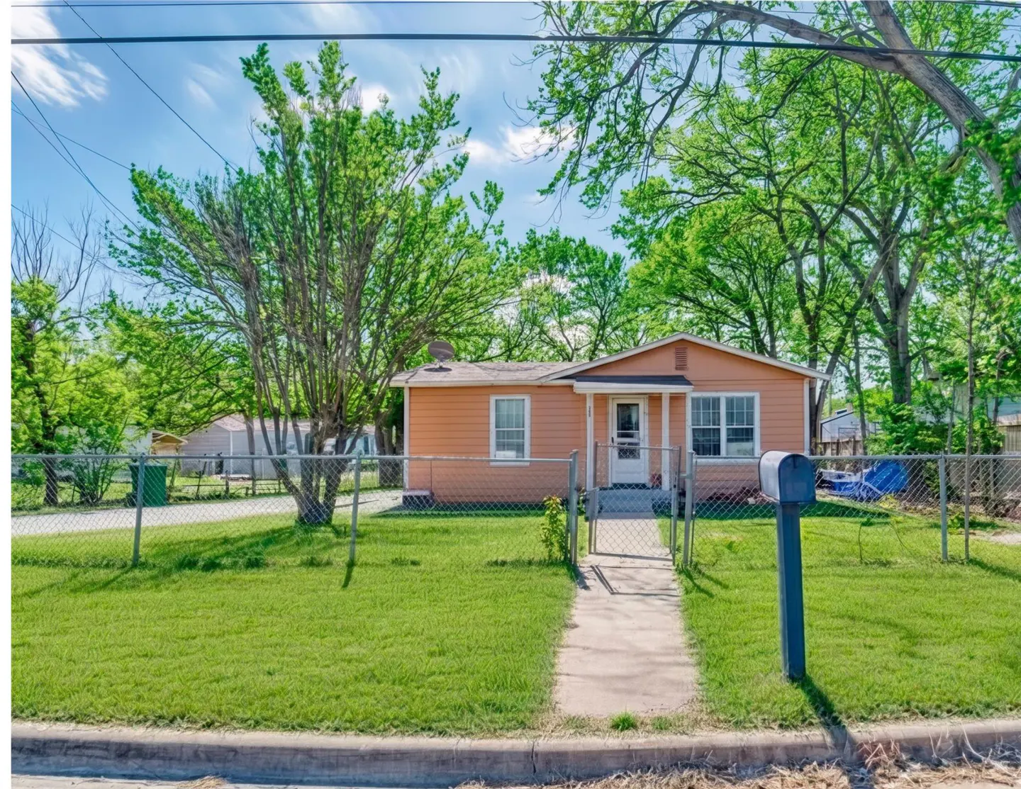 A single-story peach house with a white door and trim, surrounded by a chain-link fence and green lawn.