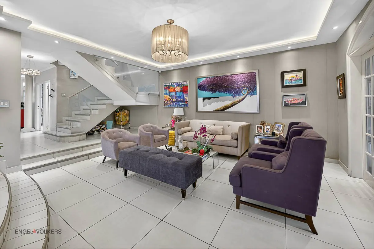 A bright living room with white tile floors, gray walls, and a staircase. Furnishings include a beige sofa, purple chairs, and colorful artwork.