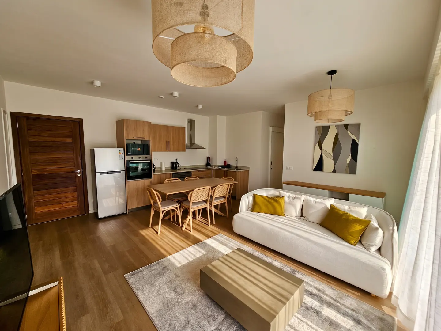 A bright, modern apartment interior with a white sofa, wooden table, and kitchen area. Sunlight streams through the window.