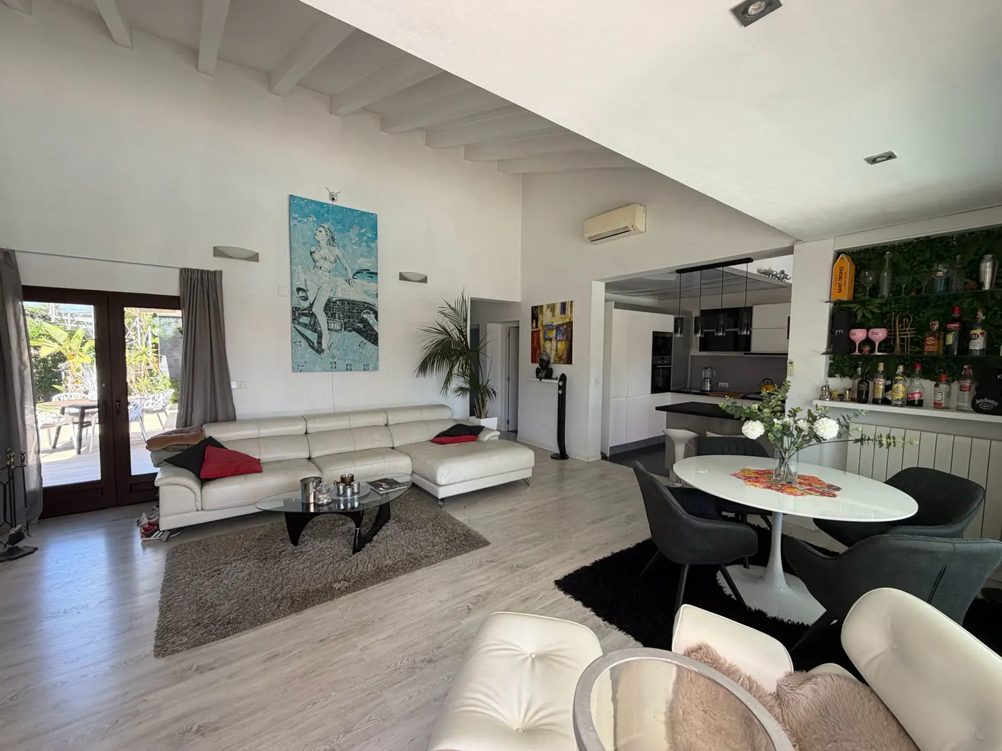 Bright, open-concept living space with white walls, light wood floors, and a high ceiling. A white sectional sofa faces a glass coffee table. Dining area and kitchen are visible.