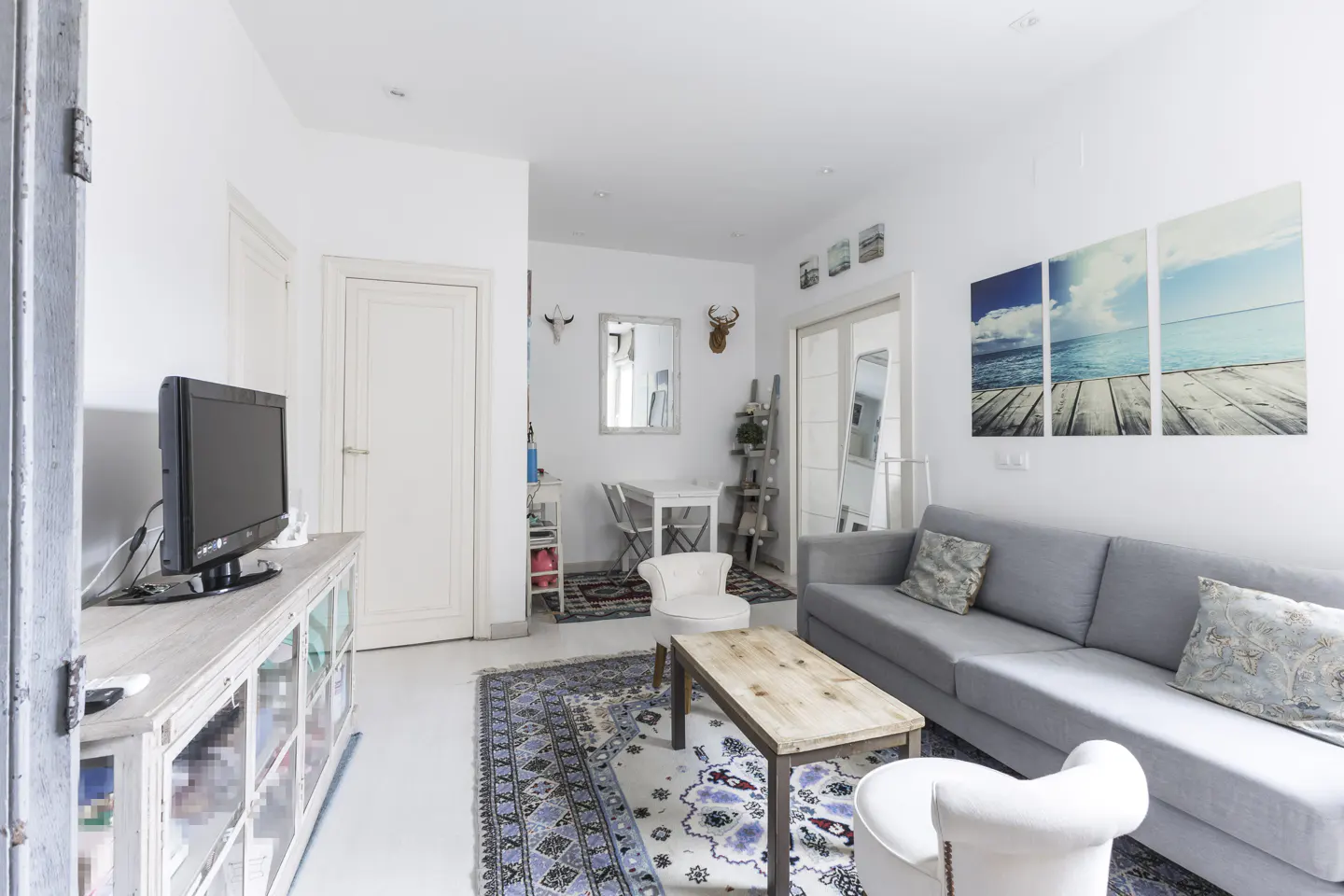 Bright living room with white walls, a gray sofa, and a wooden coffee table on a patterned rug. A triptych of a beach scene hangs above the sofa.