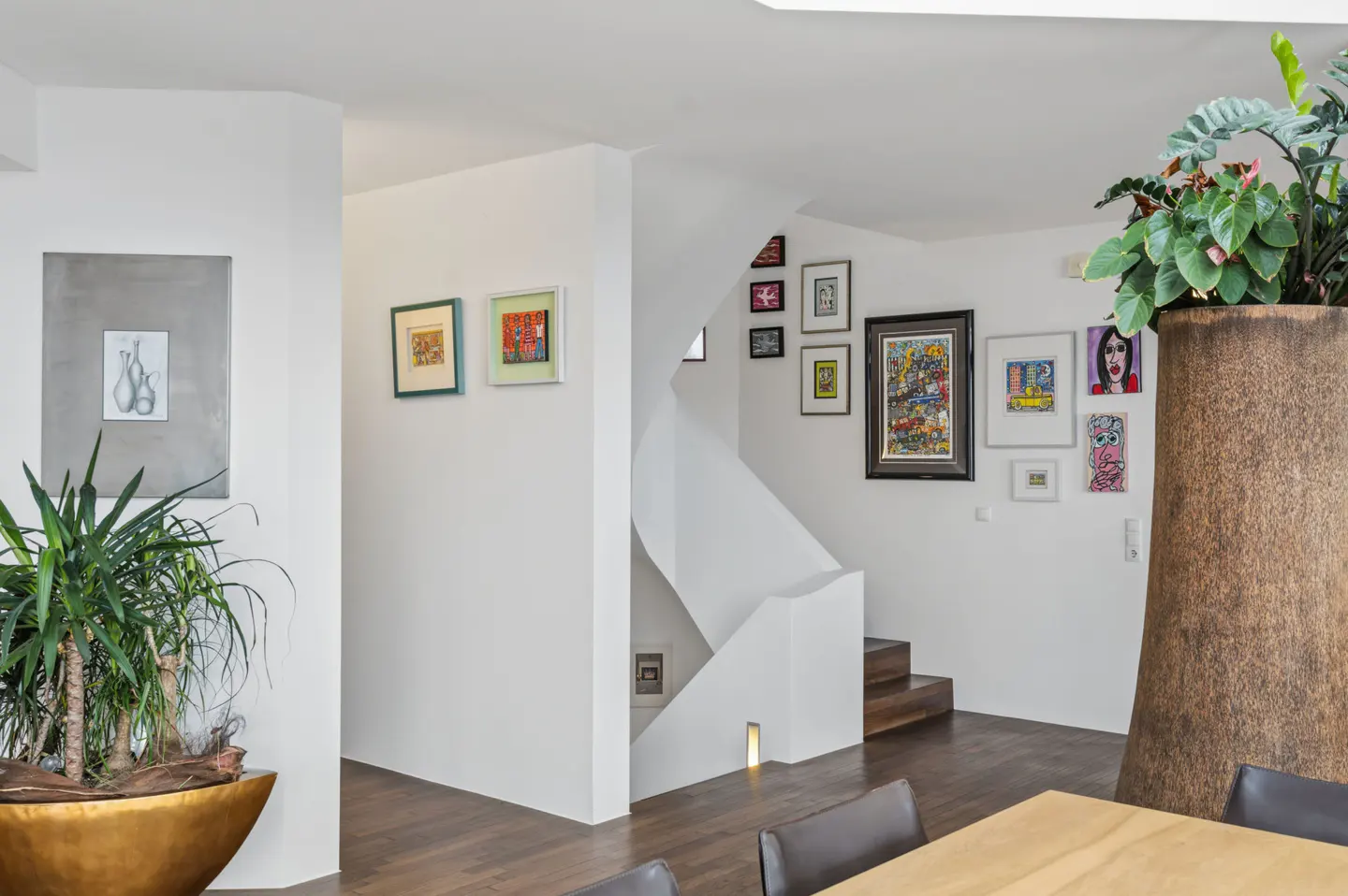 Interior view of a modern home with white walls, art, plants, and a staircase. Wood floors and a dining table are visible.