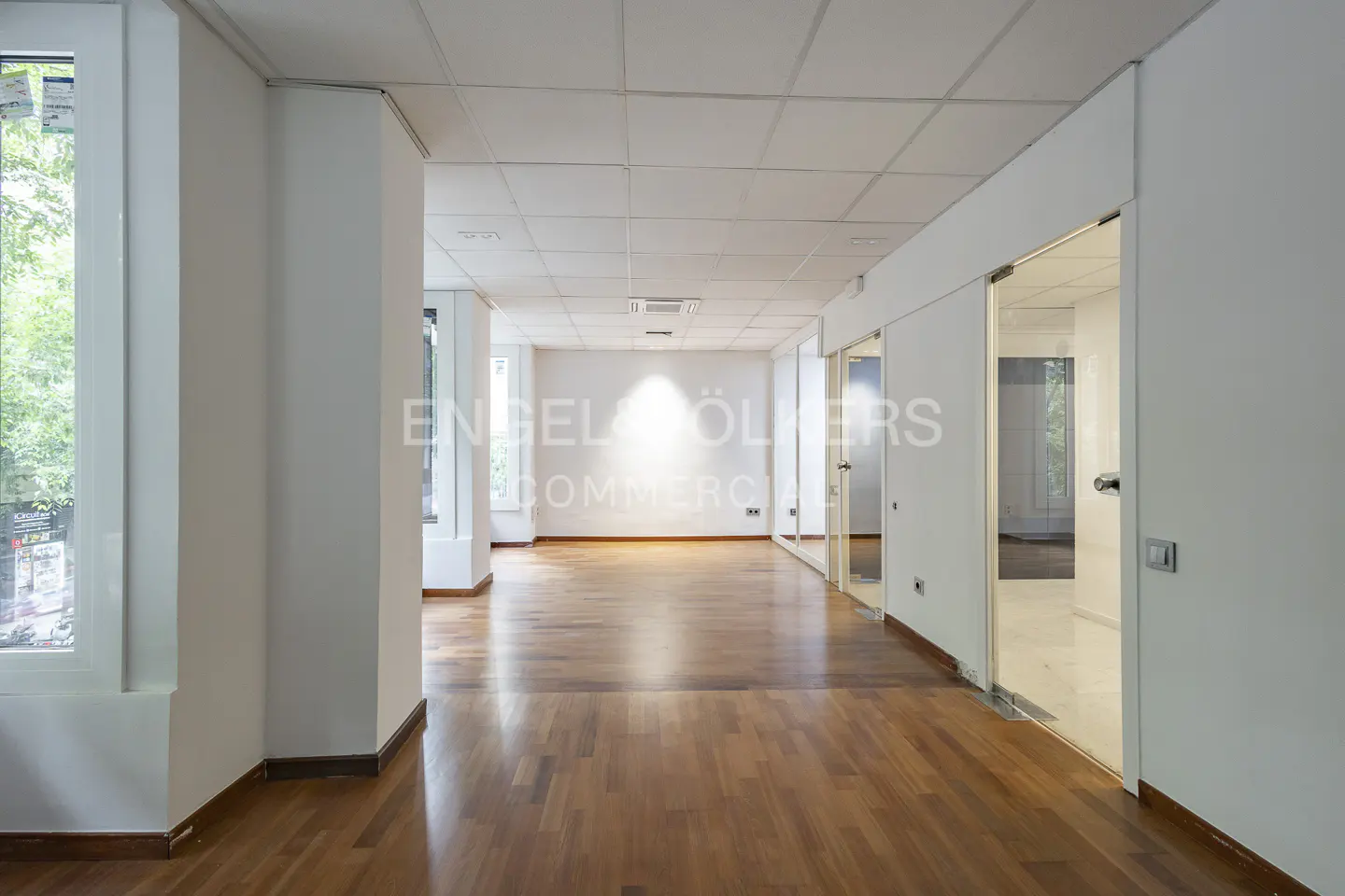 Empty office space with hardwood floors, white walls, and glass-enclosed rooms. Windows provide natural light.