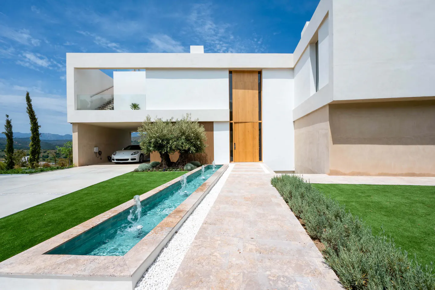 Modern white house with a long water feature leading to a wooden door. A white car is parked under the house's overhang.