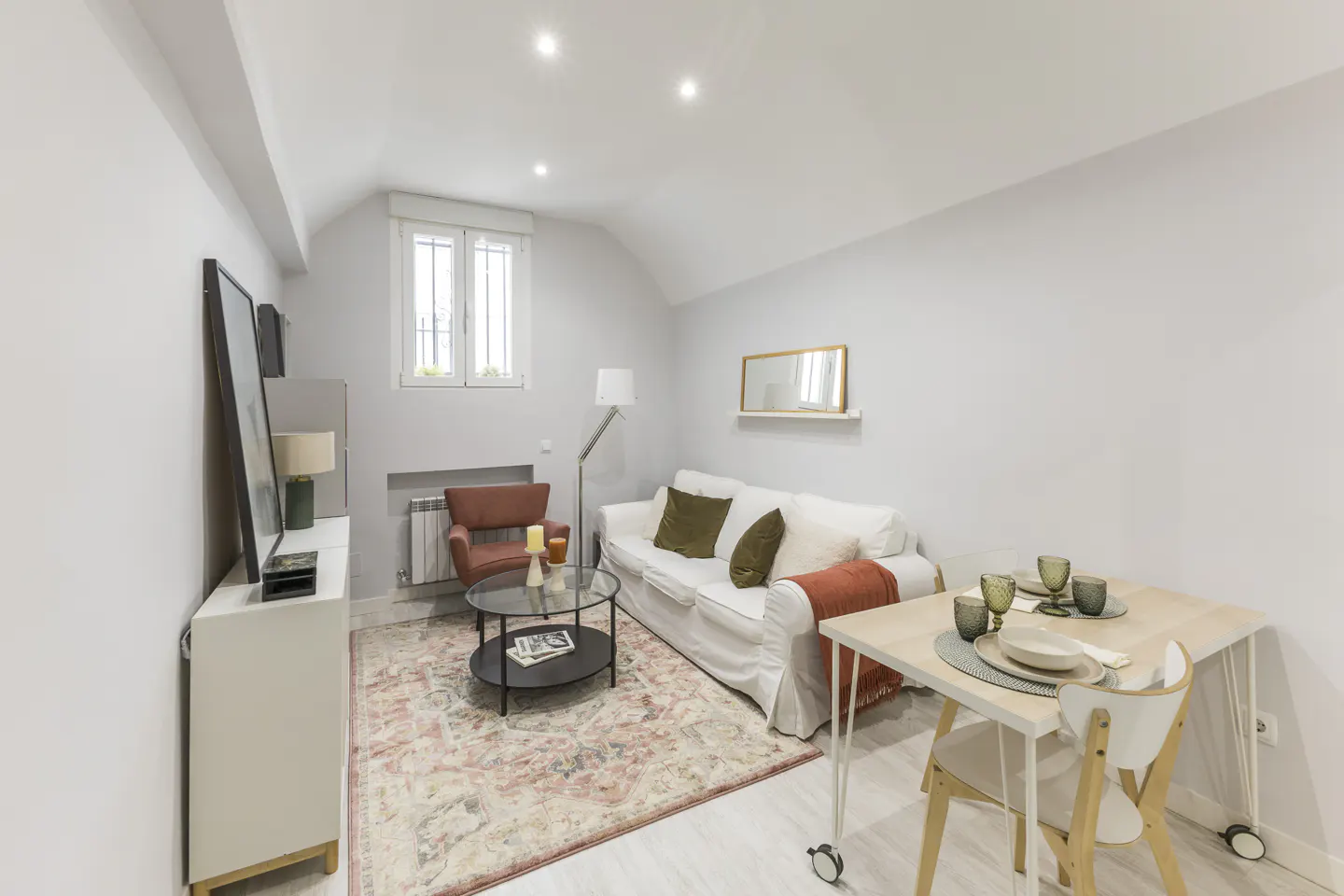 Bright living room with a white sofa, red armchair, glass coffee table, patterned rug, and a small dining area.