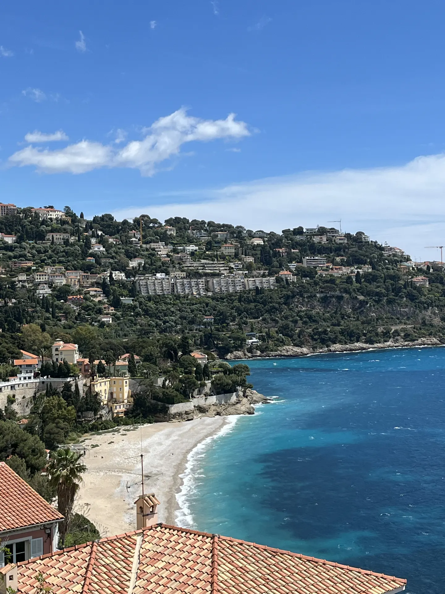 Coastal view of Villefranche-sur-Mer, France, with turquoise water, a sandy beach, and hillside villas under a blue sky.