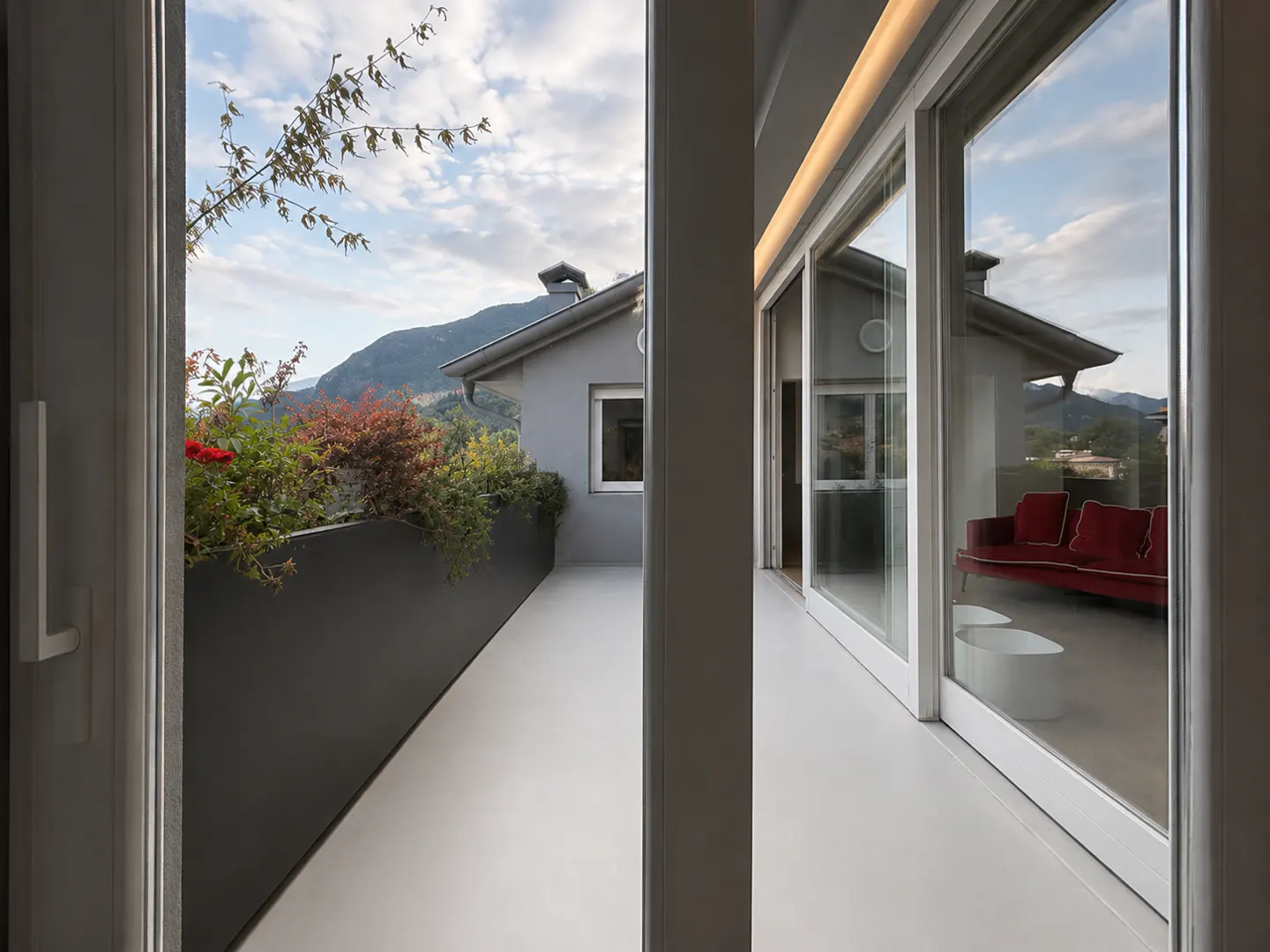 Modern balcony with a sleek grey planter, red sofa visible through glass doors, and scenic mountain views in the background.