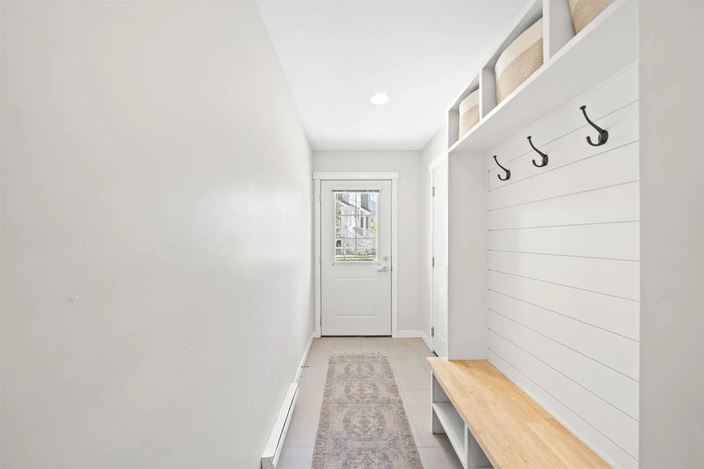 Bright entryway with white walls, a door with glass panes, and a built-in bench with hooks and storage baskets. A patterned rug runs down the center of the tiled floor.