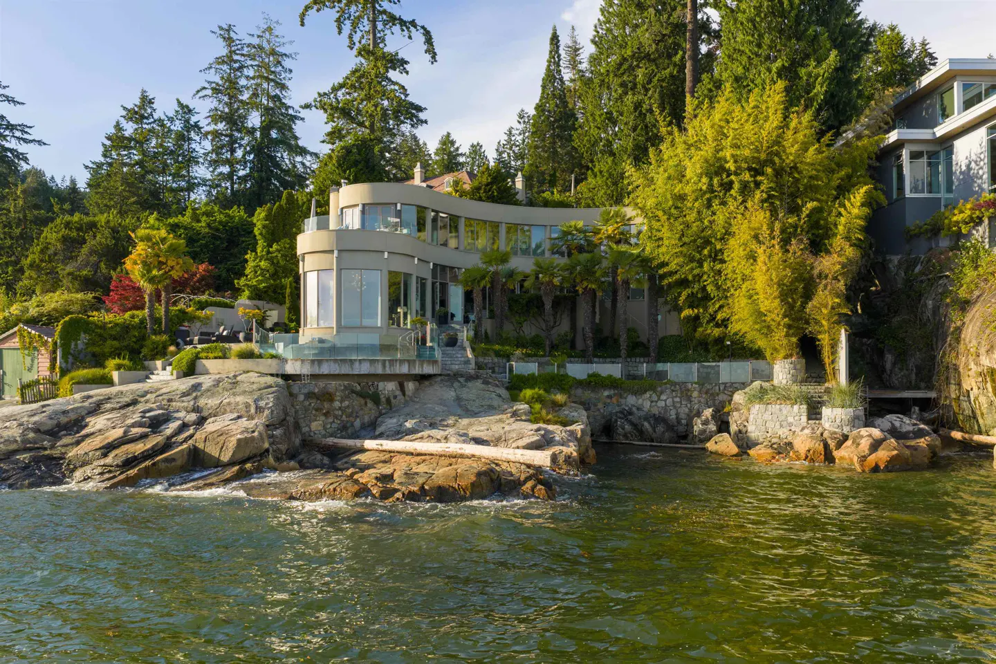 Modern curved waterfront home with glass walls, nestled on a rocky shore surrounded by lush trees and greenery.