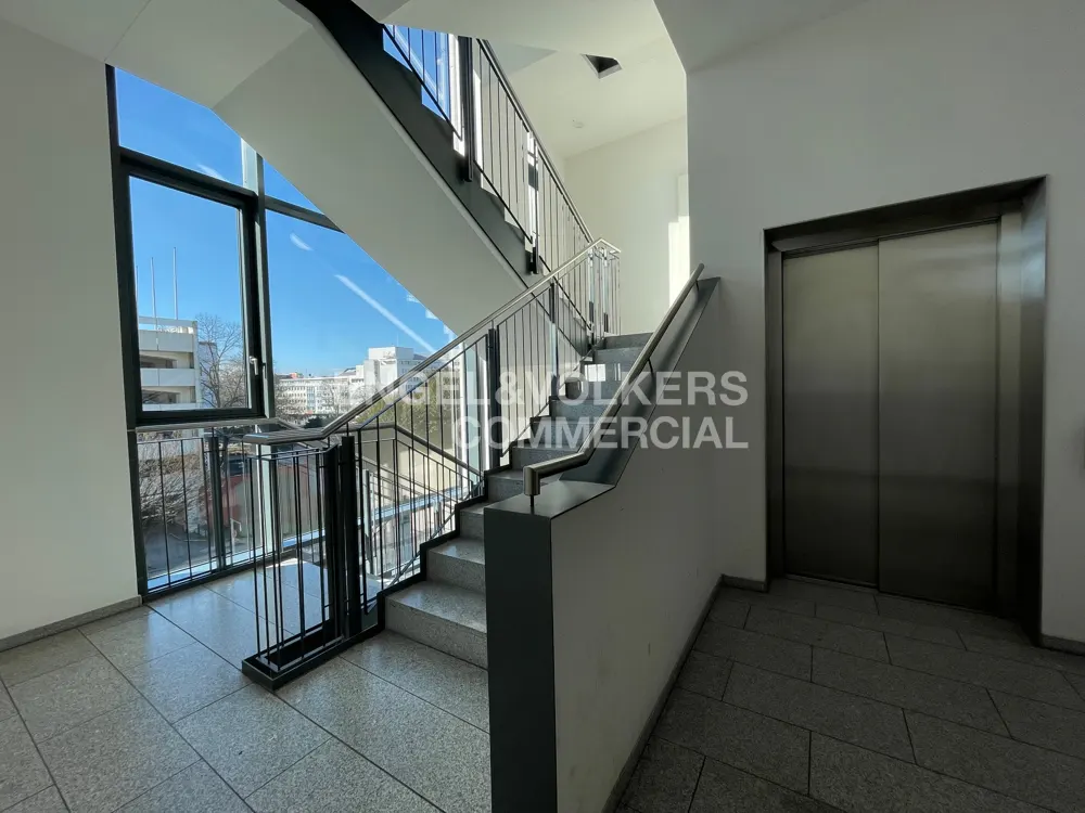 Interior view of a building with stairs, a metal elevator, and a large window showing a city view.