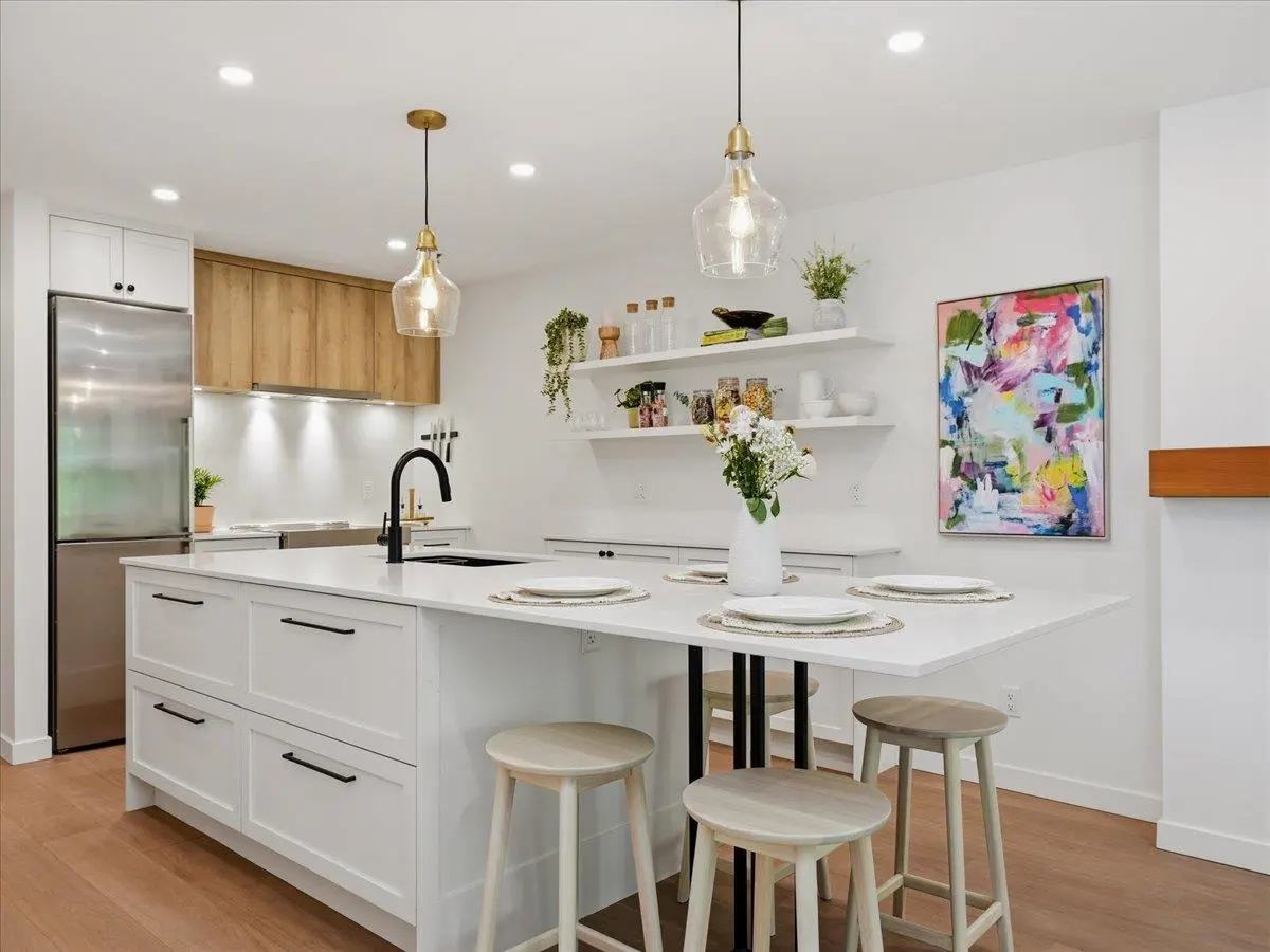 Bright, modern kitchen with white cabinets, stainless steel fridge, and wood floors. Island has seating and pendant lights hang above. Shelves display plants and decor.