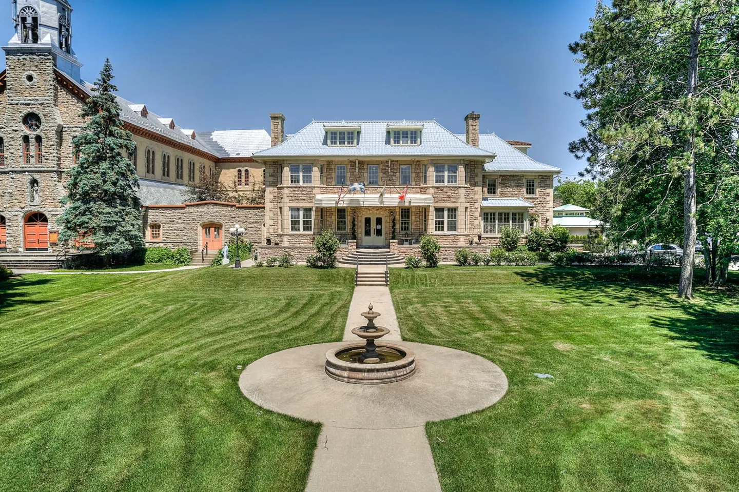 Stone building with a fountain in front. The building has a blue roof and a green lawn. There are trees on either side of the building.