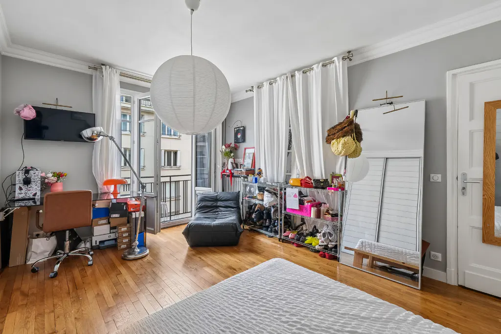 Bright bedroom with hardwood floors, white walls, and a balcony. A large paper lantern hangs from the ceiling. A desk, chair, and shoe rack are visible.
