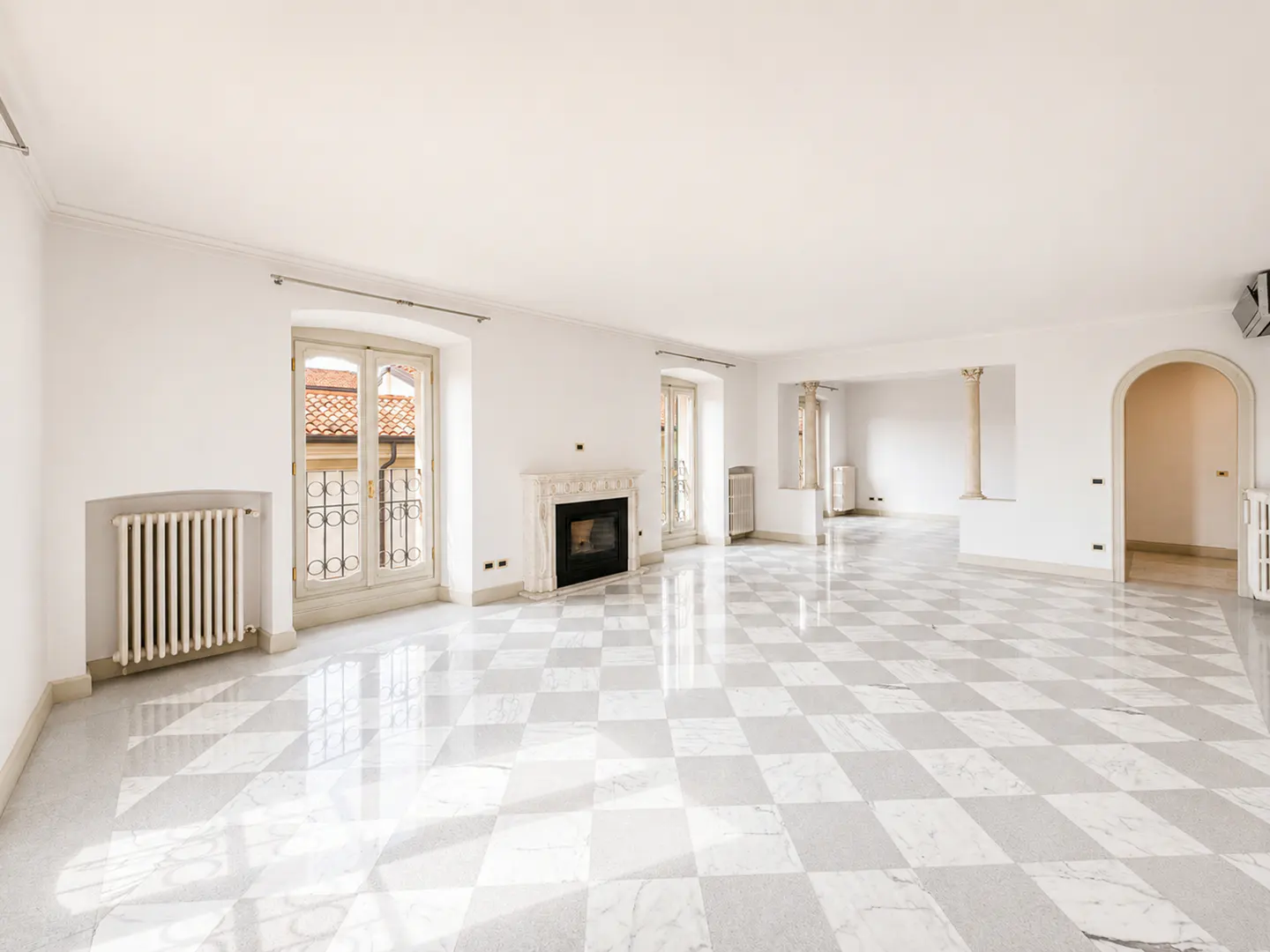 Bright, empty room with white walls, a marble fireplace, and a checkered marble floor. Windows with iron railings are visible.
