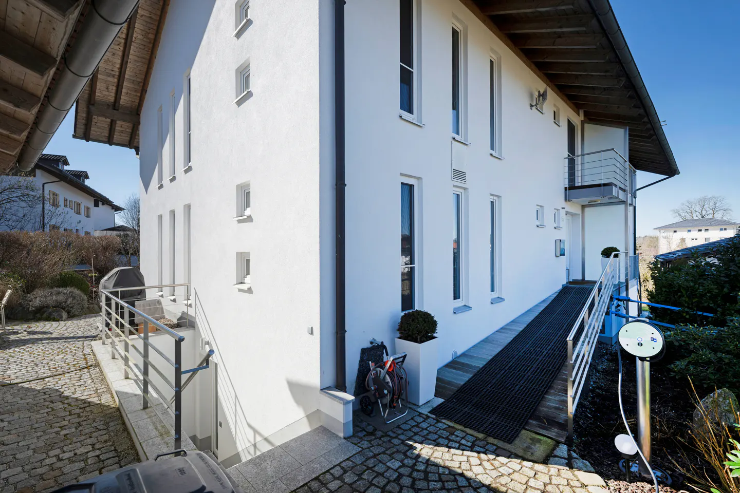 Exterior view of a modern white house with a ramp, balcony, and cobblestone driveway. A bicycle and garden equipment are visible.