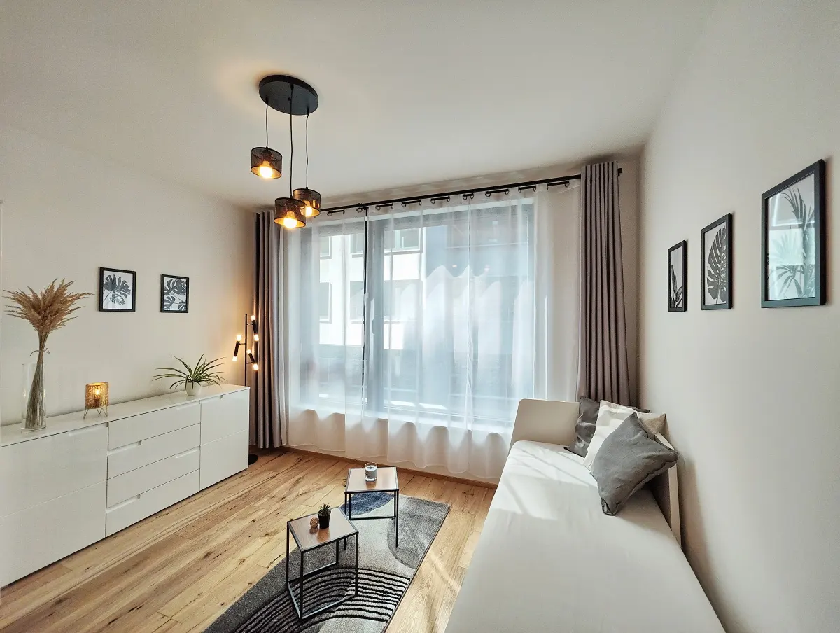 Bright living room with wood floors, white sofa, and a large window with sheer curtains. Black framed art hangs on the wall.
