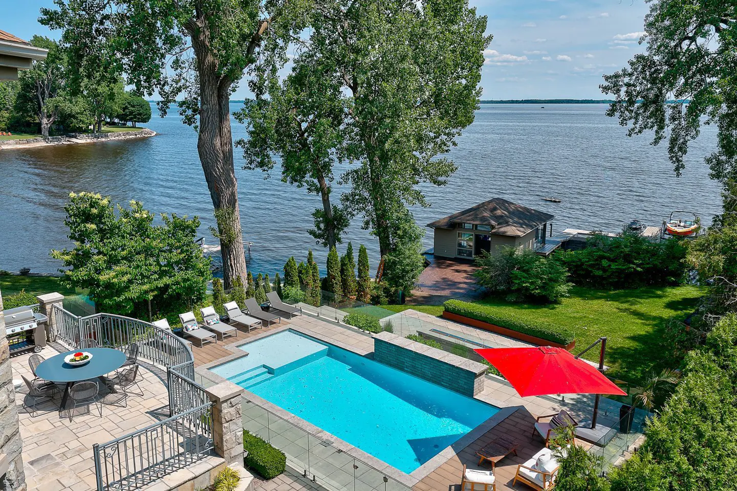 Aerial view of a backyard with a pool, patio, and lake access. The pool is surrounded by lounge chairs and a red umbrella. A boathouse sits on the lake.