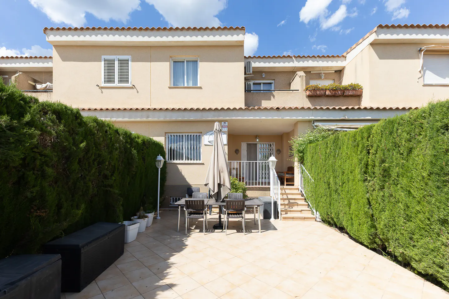Backyard patio of a beige two-story townhouse with a table, chairs, and green hedges.