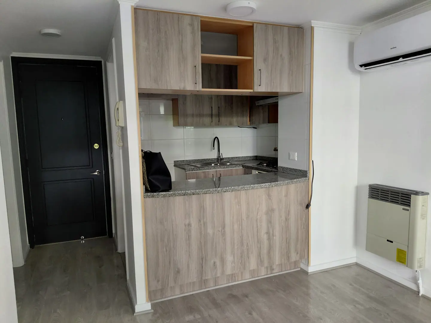A bright, modern apartment interior with a kitchen area featuring wood-grain cabinets, granite countertops, and a black door.