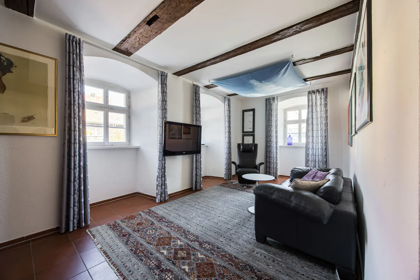 Living room with exposed beams, white walls, and a patterned rug. A black leather sofa and armchair provide seating. Windows have patterned curtains.