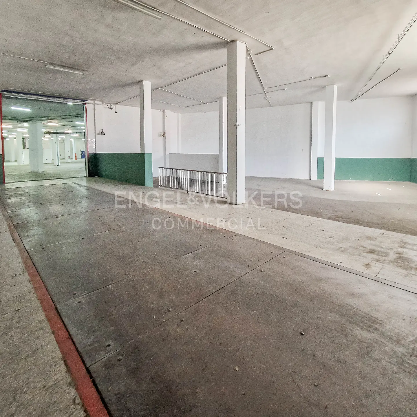 Empty commercial space with concrete floors, white walls, and support columns. A loading dock is visible in the background.
