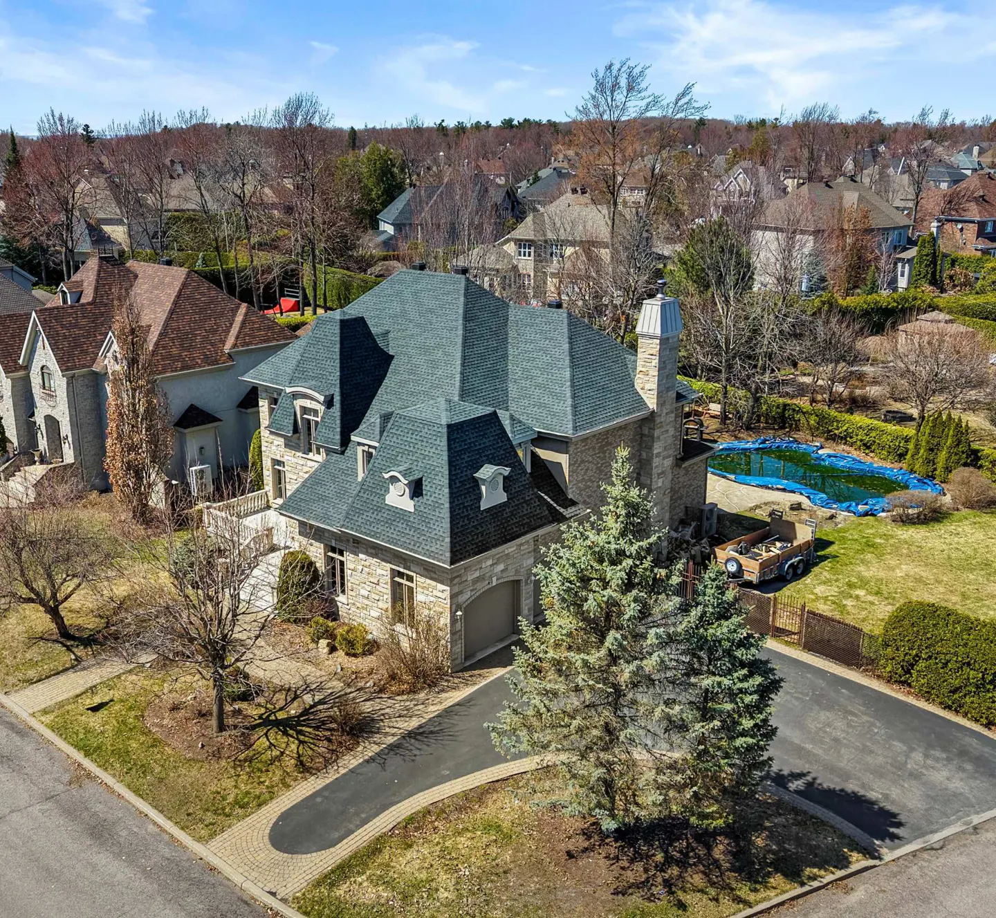 Aerial view of a large stone house with a dark green roof, a pool covered with a blue tarp, and a paved driveway.