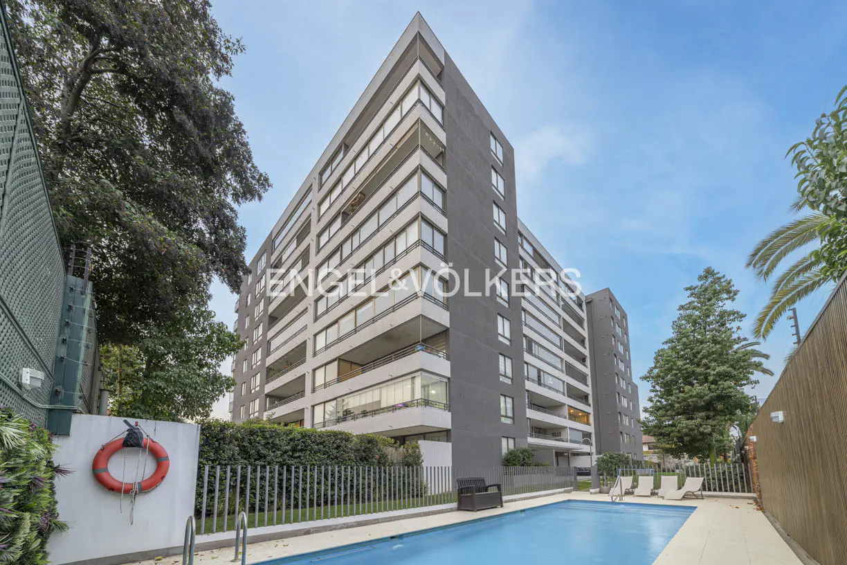 Modern apartment building with balconies overlooking a swimming pool and lounge chairs under a clear blue sky.
