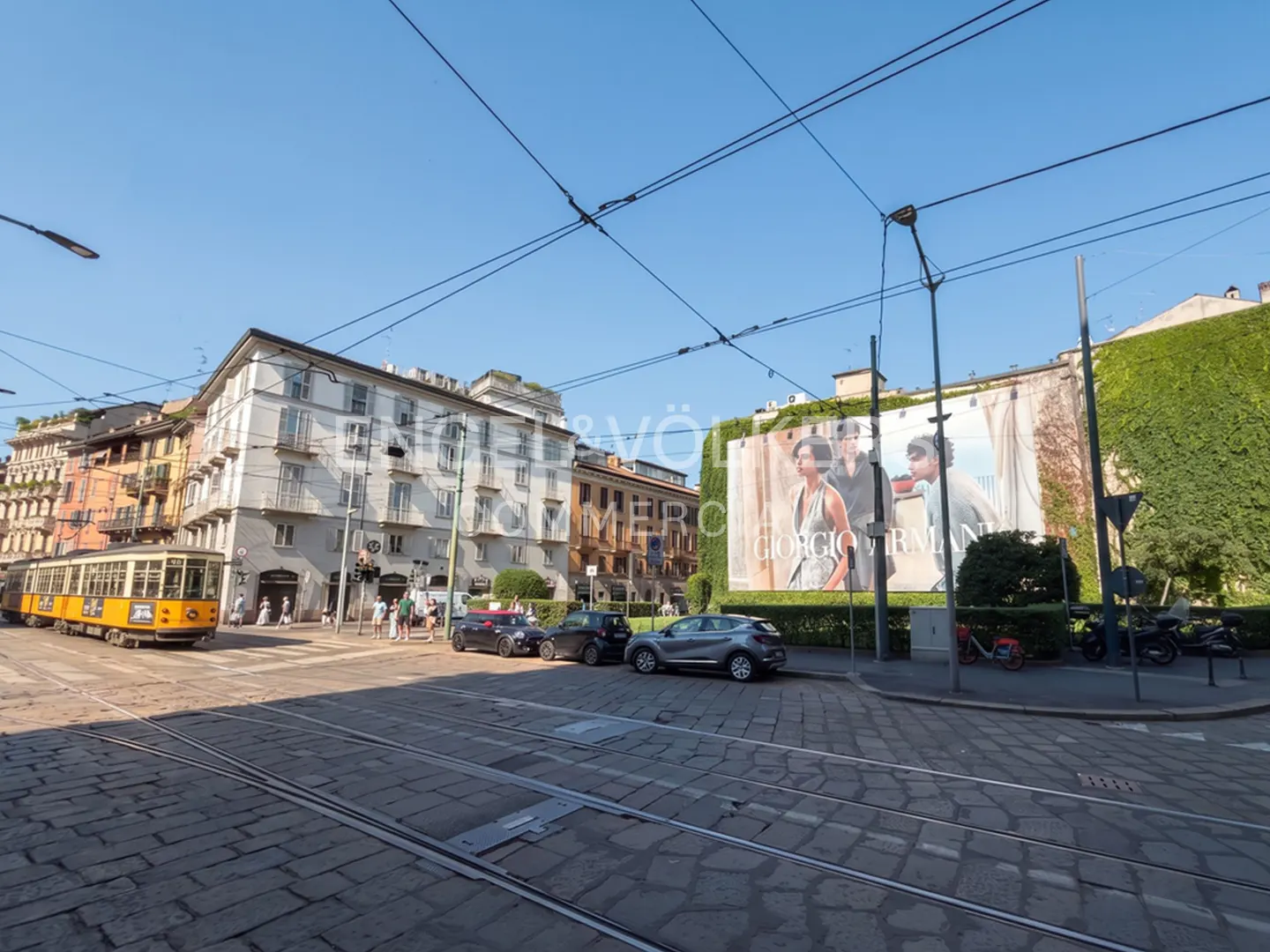 A yellow tram travels along tracks on a cobblestone street in Milan, past parked cars and a large Giorgio Armani billboard.