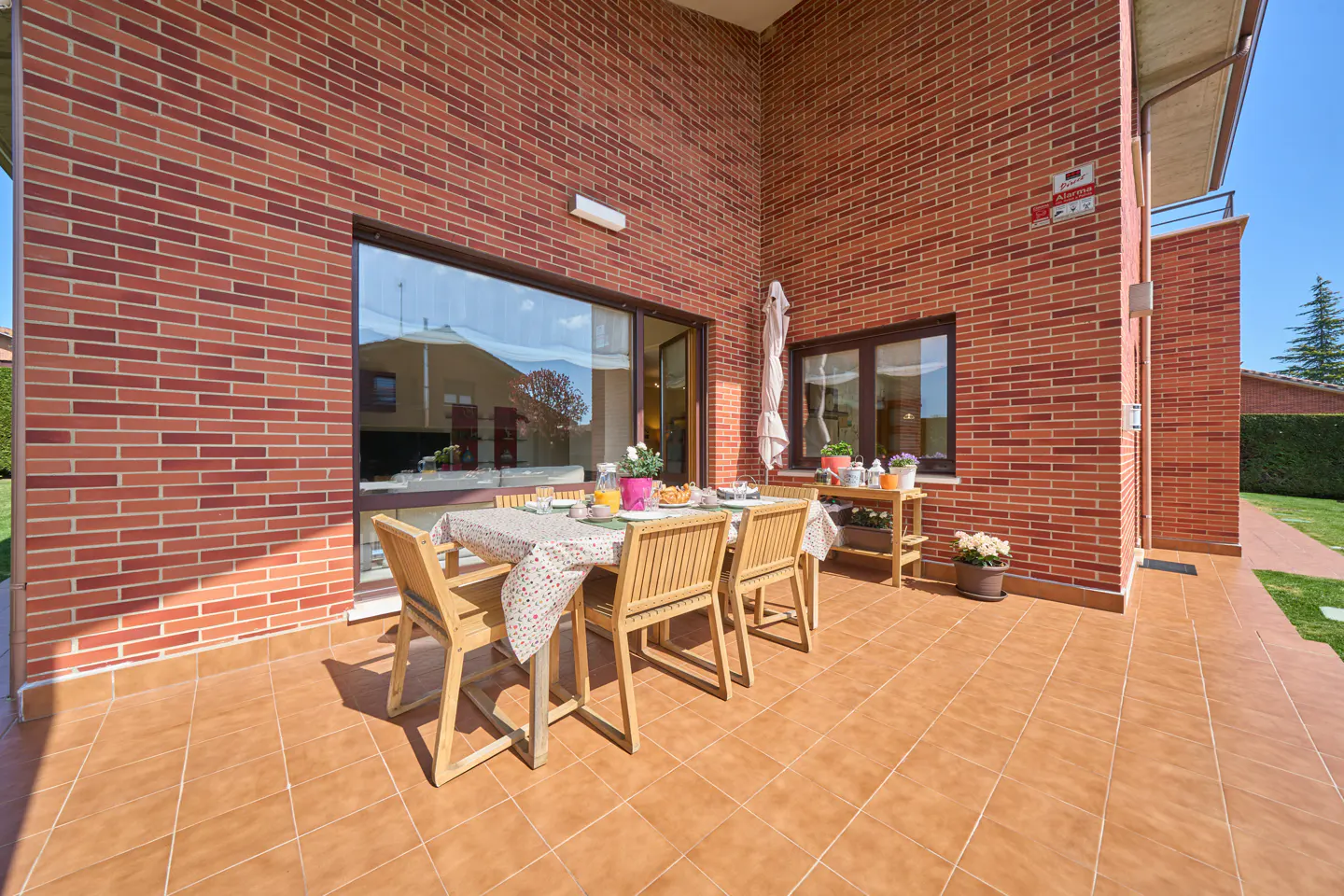 Outdoor patio with a brick wall, brown tile floor, and a wooden table set for a meal. A small wooden table with flowers sits nearby.