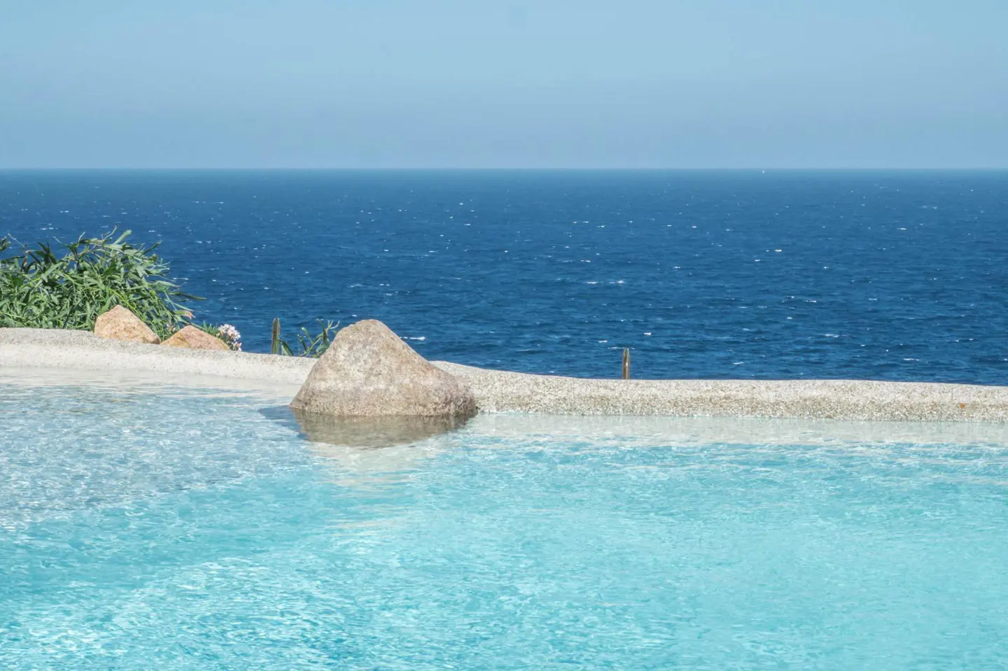 Infinity pool with a large stone at the edge, overlooking the deep blue ocean under a clear sky.