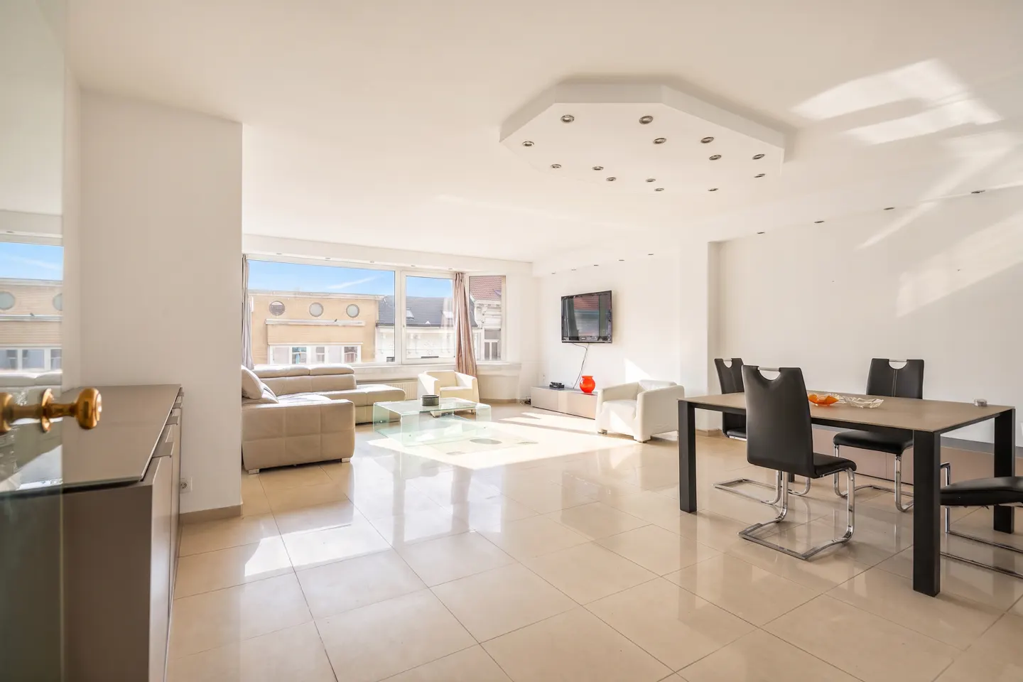 Bright, open-plan living and dining area with beige tile floors, white walls, and large windows. Beige sofa, white chairs, and a black dining table with chairs.