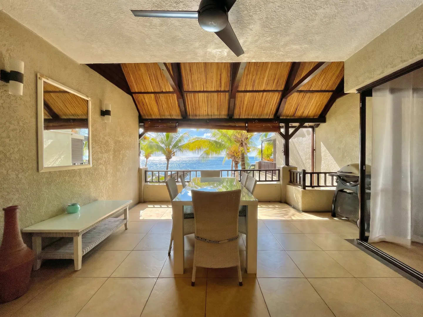 Covered patio with a dining table and ocean view. The patio has a thatched roof, a ceiling fan, and a white table with six chairs. Palm trees and the ocean are visible in the background.