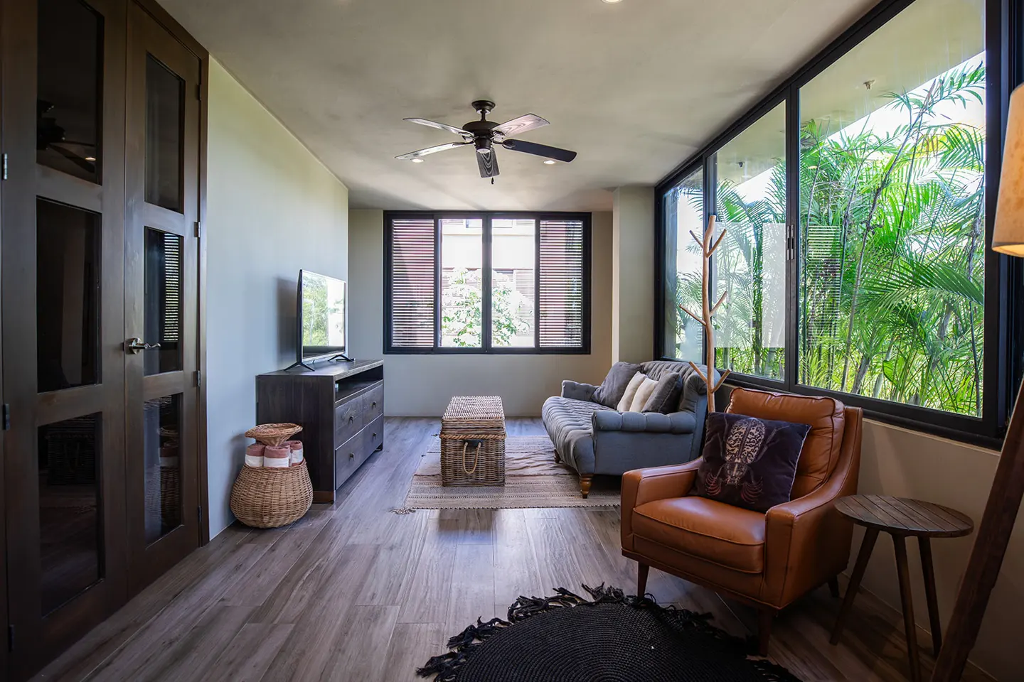 Living room with wood floors, a blue sofa, a leather chair, and large windows with a view of lush greenery.