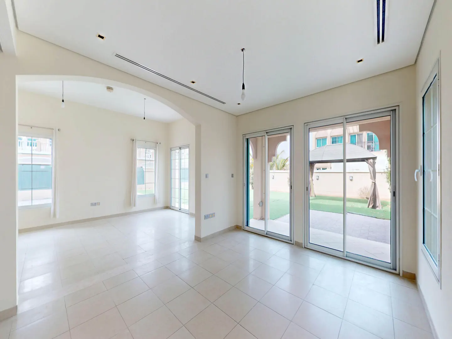 Bright, empty room with white walls, tile floor, and large windows. Sliding glass doors lead to a green lawn and outdoor gazebo.