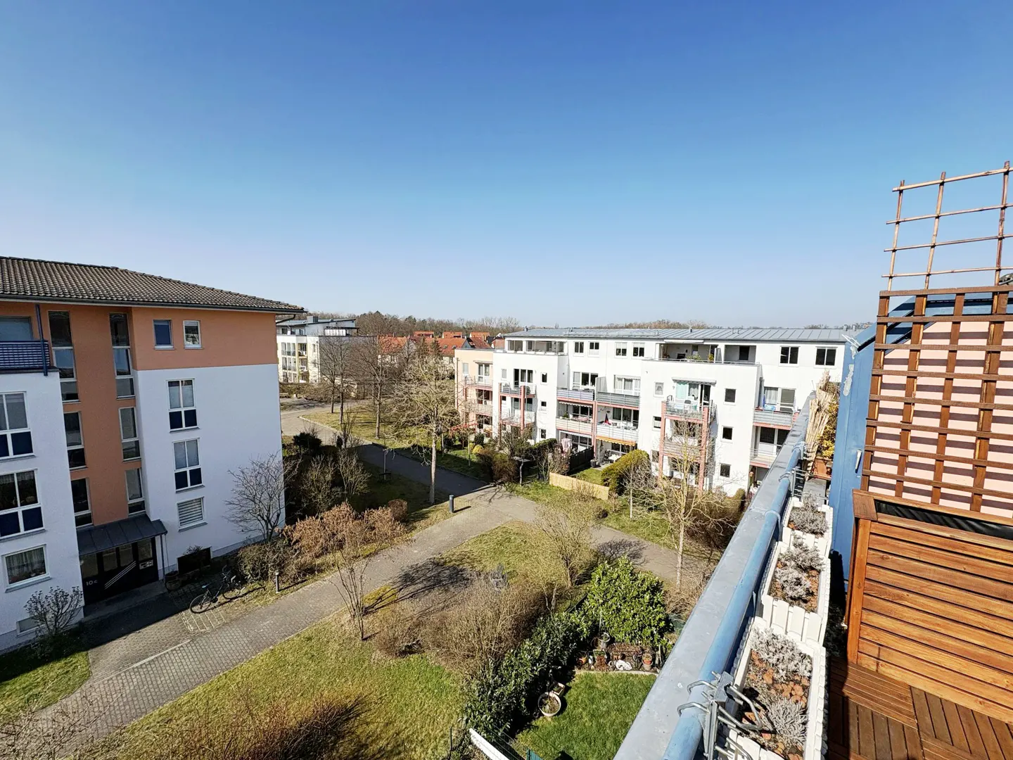 View from a balcony overlooking apartment buildings, green spaces, and a clear blue sky. Balcony has wooden privacy screen and potted plants.