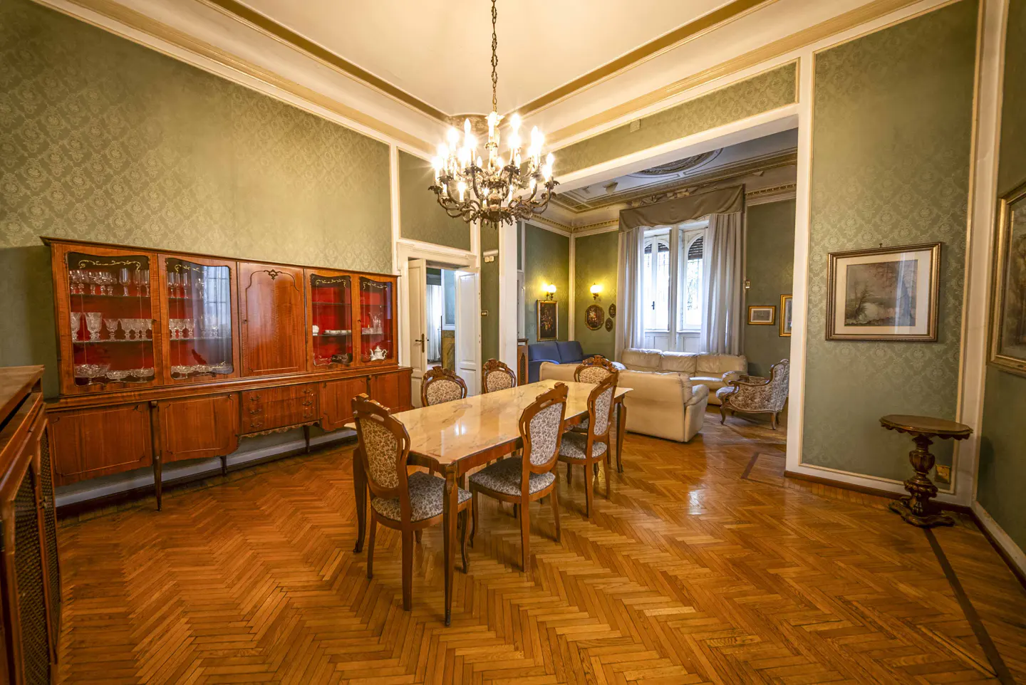 A dining room with a wooden table and chairs, a large wooden cabinet, and a chandelier. The walls are green and the floor is herringbone wood.