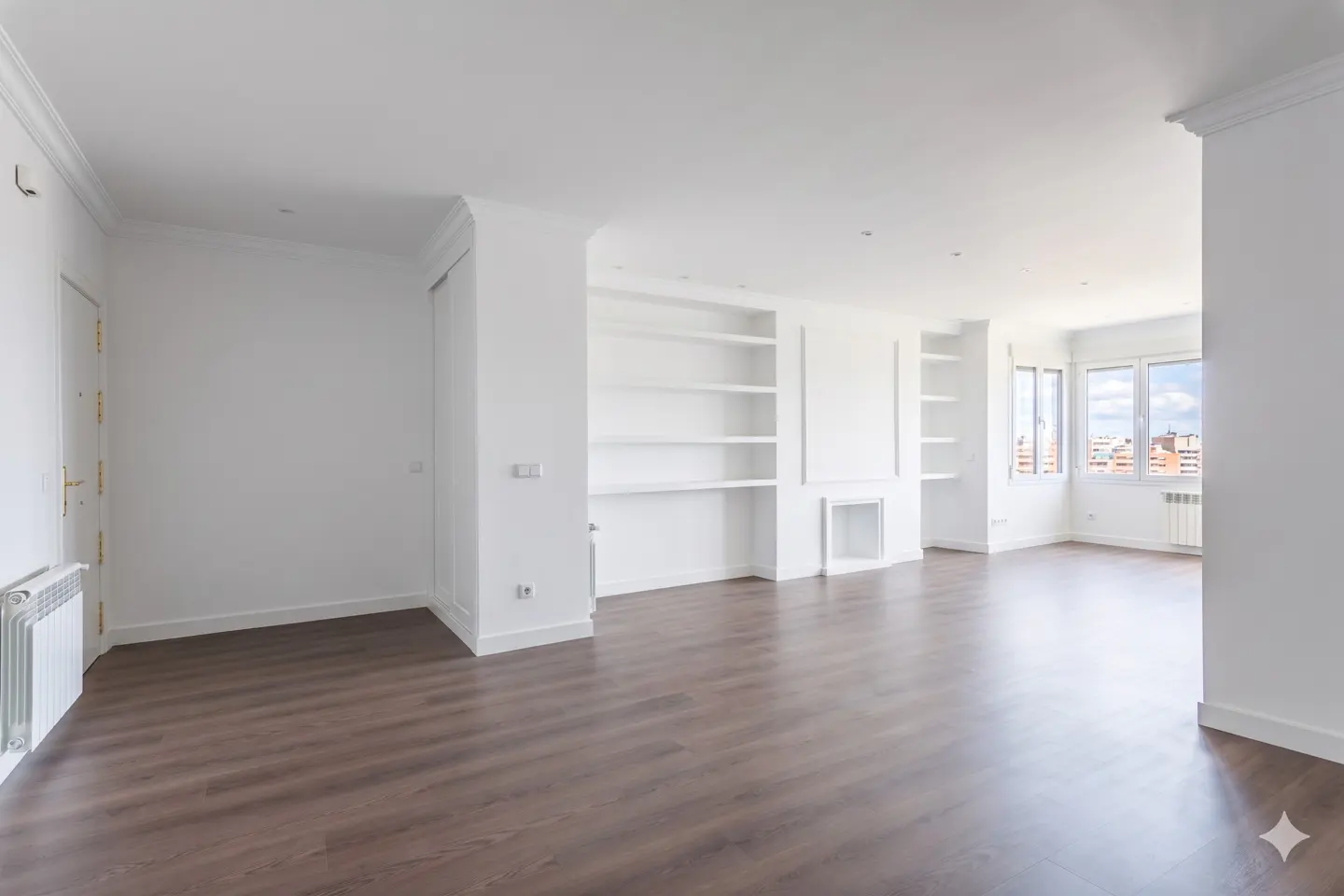 Bright, empty room with dark wood floors and white walls. Built-in white shelves and a window with a city view.