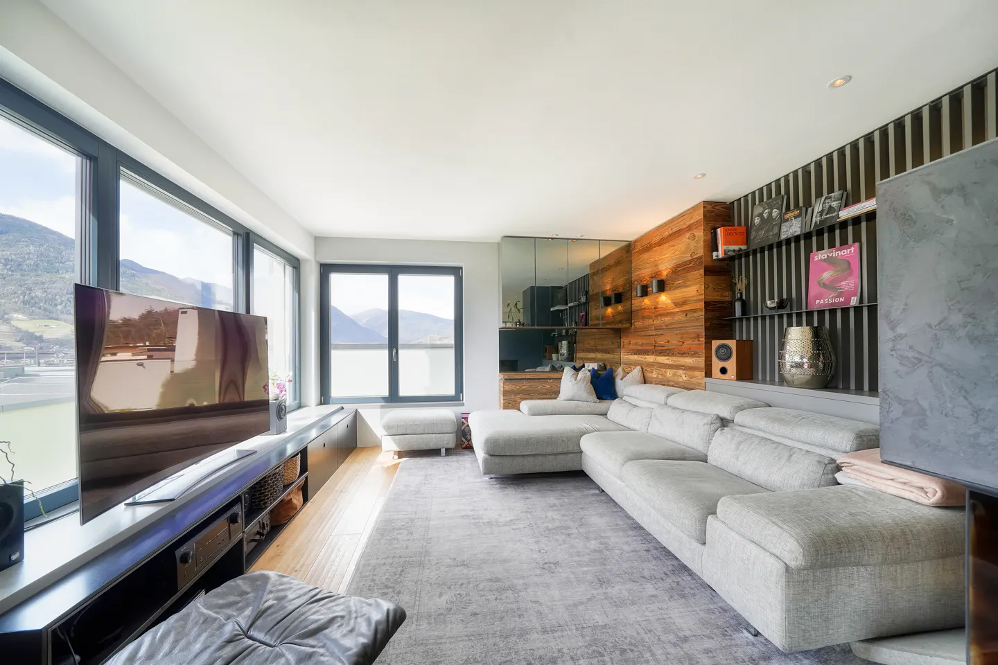 Bright living room with a gray sectional sofa, large windows with mountain views, and a wood-paneled wall with shelves.