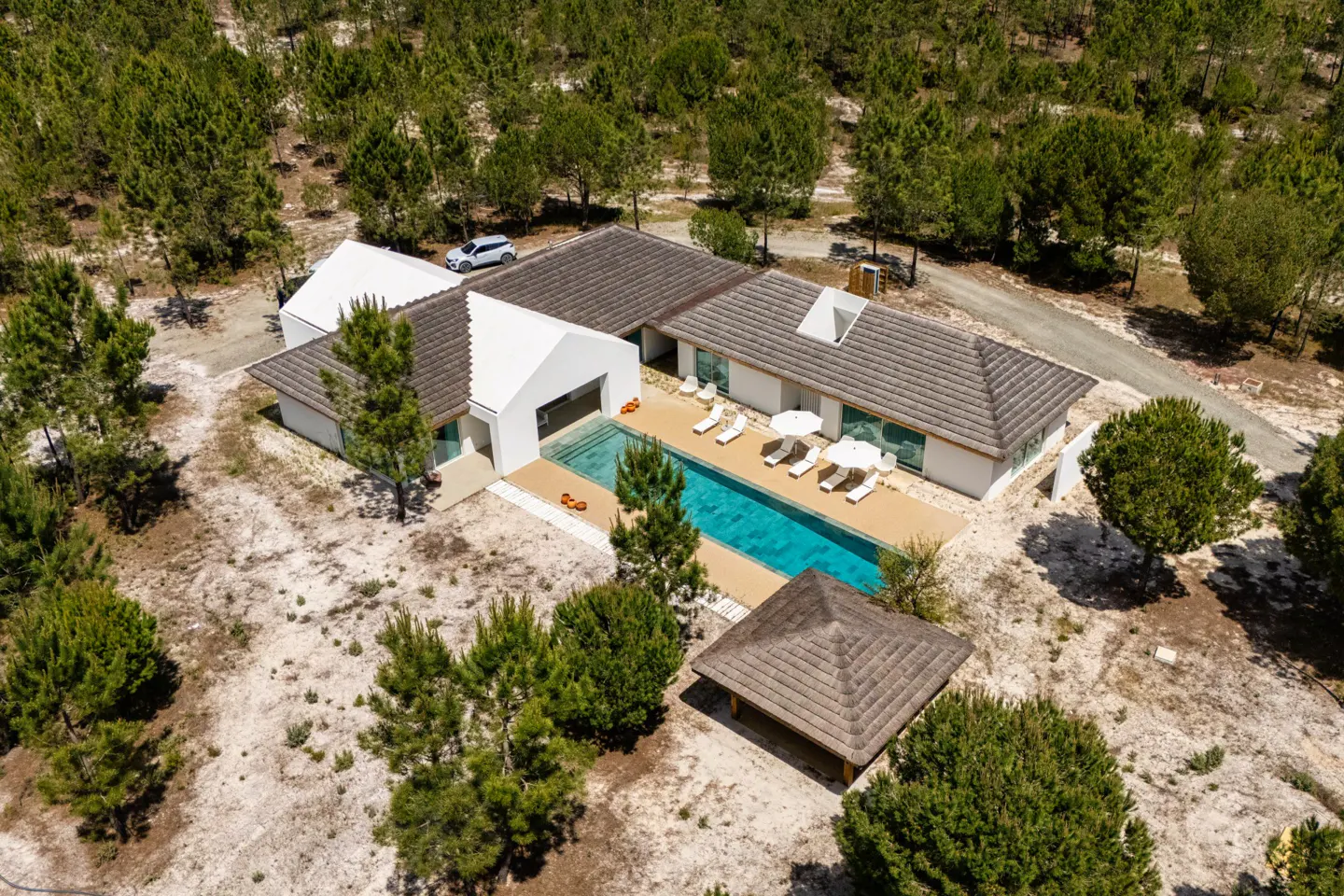 Aerial view of a modern white house with a pool, lounge chairs, and a gazebo surrounded by trees.
