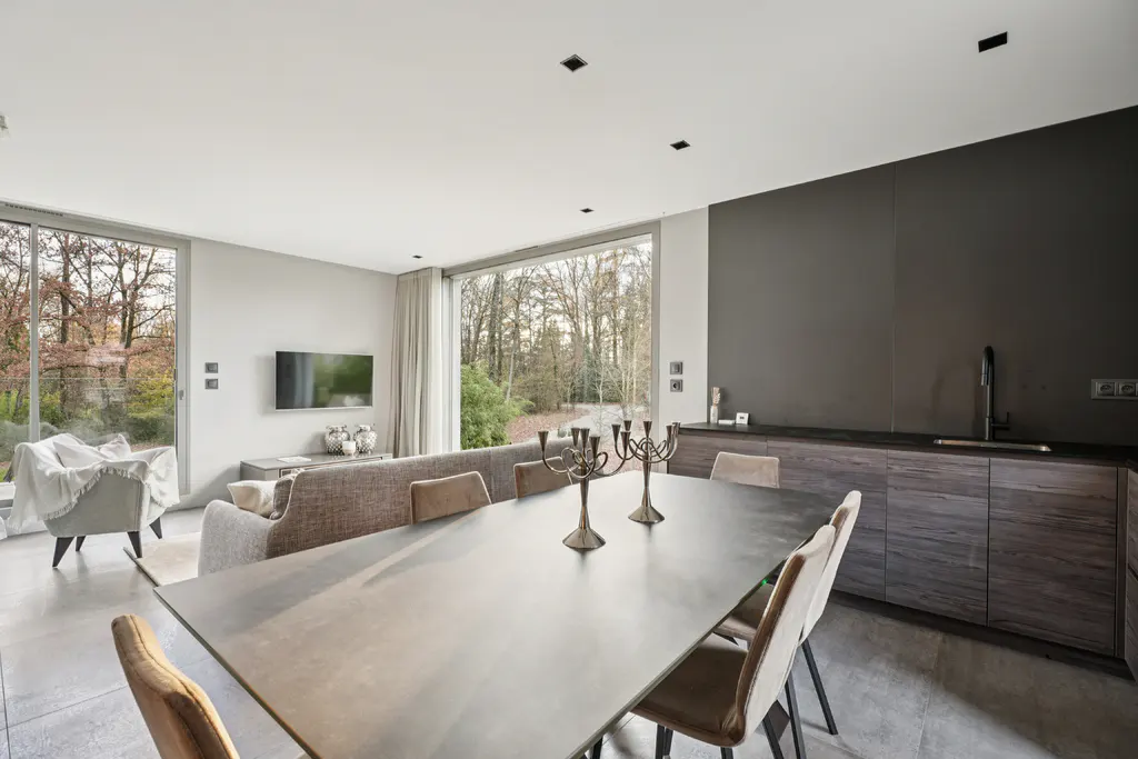 A modern open-concept living space with a dining table, chairs, and a view of trees through large windows.